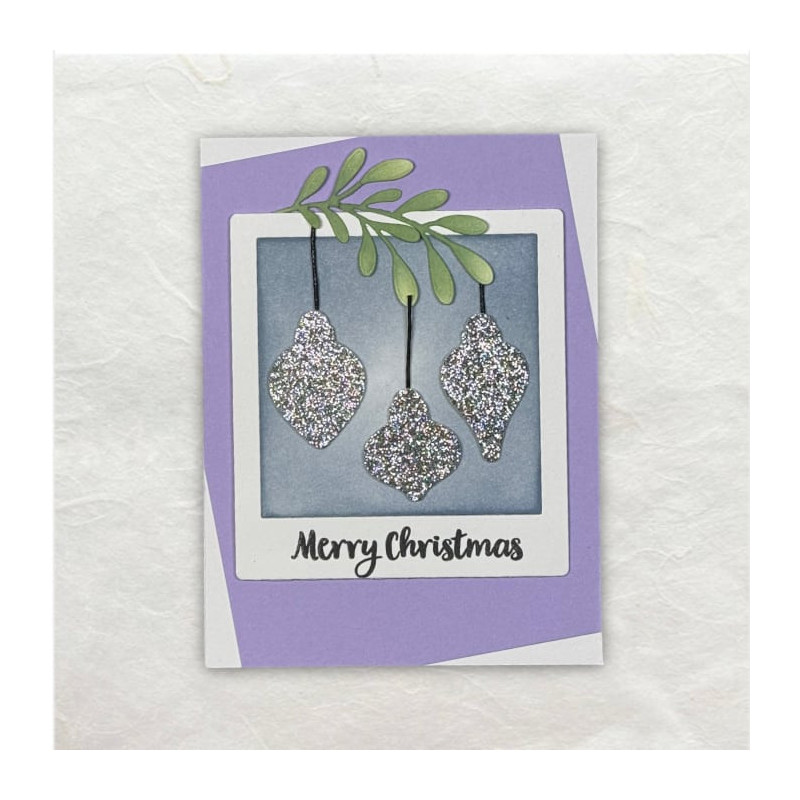 Glitterende gestanste Kerstballen in een soort Polaroid frame in lila hangend aan een groen takje.