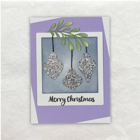 Glitterende gestanste Kerstballen in een soort Polaroid frame in lila hangend aan een groen takje.